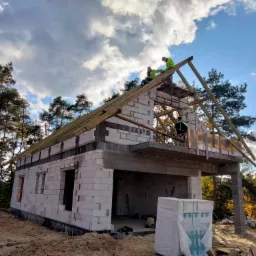 Wznoszenie budynku z białych bloczków, widoczni pracownicy na dachu montujący konstrukcję drewnianą, w tle las i błękitne niebo z chmurami.