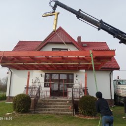 DREW-DOM Andrzej Antecki - Montaż drewnianej pergoli przy domu jednorodzinnym z użyciem dźwigu, widoczni pracownicy w odzieży roboczej, elementy konstrukcyjne w kolorze czerwonym.