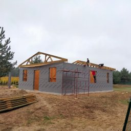 DREW-DOM Andrzej Antecki - Wznoszenie konstrukcji domu z drewnianym szkieletem; widoczne rusztowania, izolacja zewnętrzna STROTEX, ekipa montażowa na dachu, stos drewna na pierwszym planie i sosna po lewej stronie.