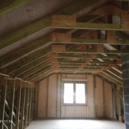 DREW-DOM Andrzej Antecki - Wnętrze poddasza w trakcie budowy z widoczną konstrukcją szkieletową, oknem i kominem z bloczków betonowych.