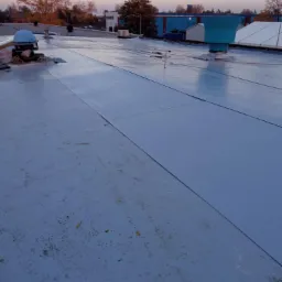 Szczelnie pokryty, płaski dach z jasnoszarej membrany bitumicznej, widoczne detale łączeń, wentylatory dachowe i elementy mocujące, w tle budynki i drzewa o zmierzchu.