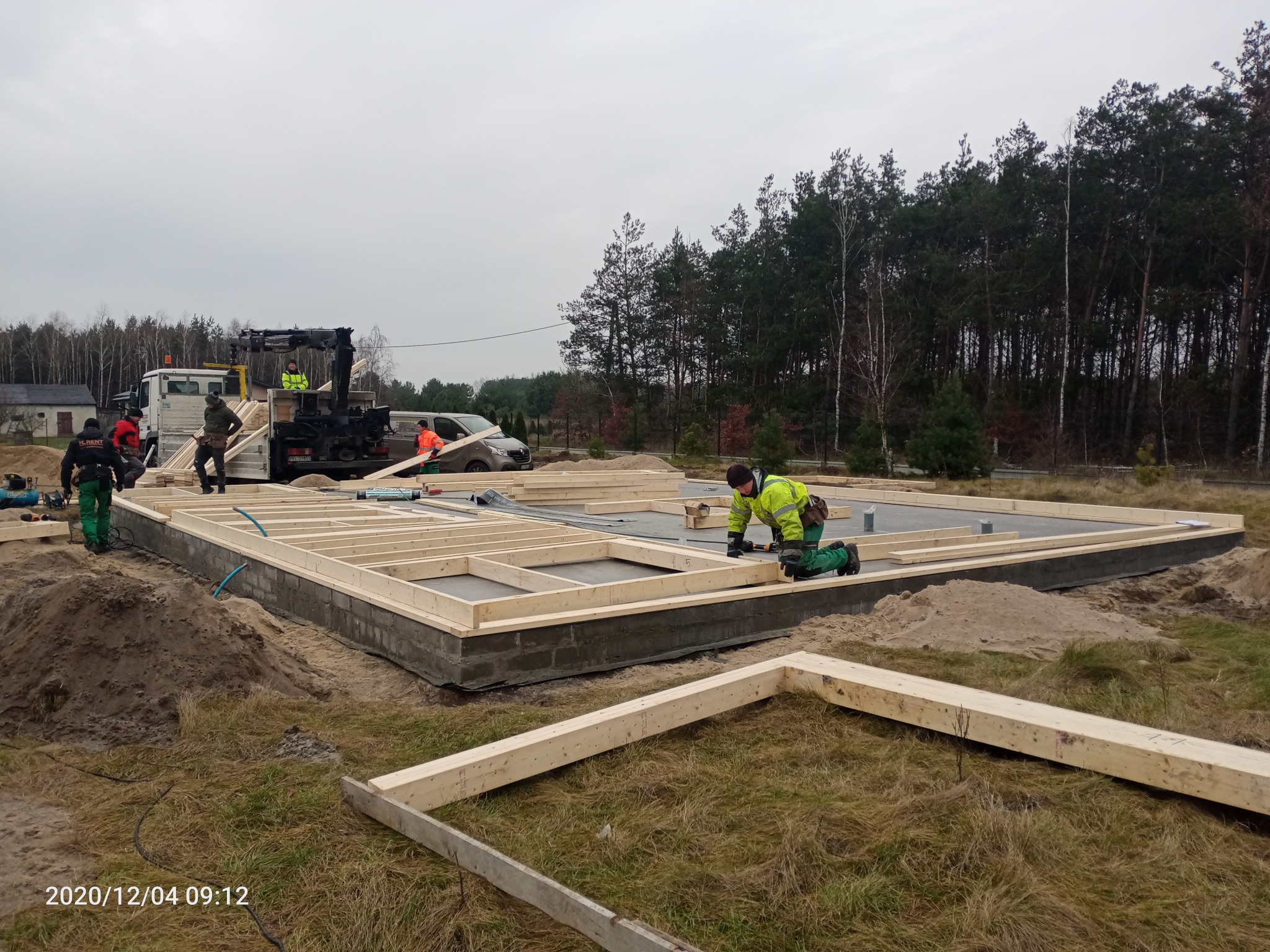 Montaż drewnianej konstrukcji fundamentu pod budynek, widoczni pracownicy w odblaskowych ubraniach i dźwig na samochodzie ciężarowym w tle.