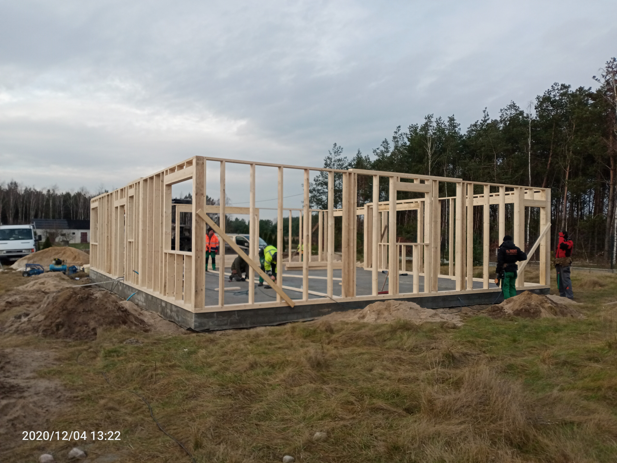 Wznoszenie drewnianej konstrukcji domu w systemie szkieletowym na betonowej płycie fundamentowej; widoczni pracownicy w odblaskowych ubraniach, sprzęt budowlany i sterta piasku.