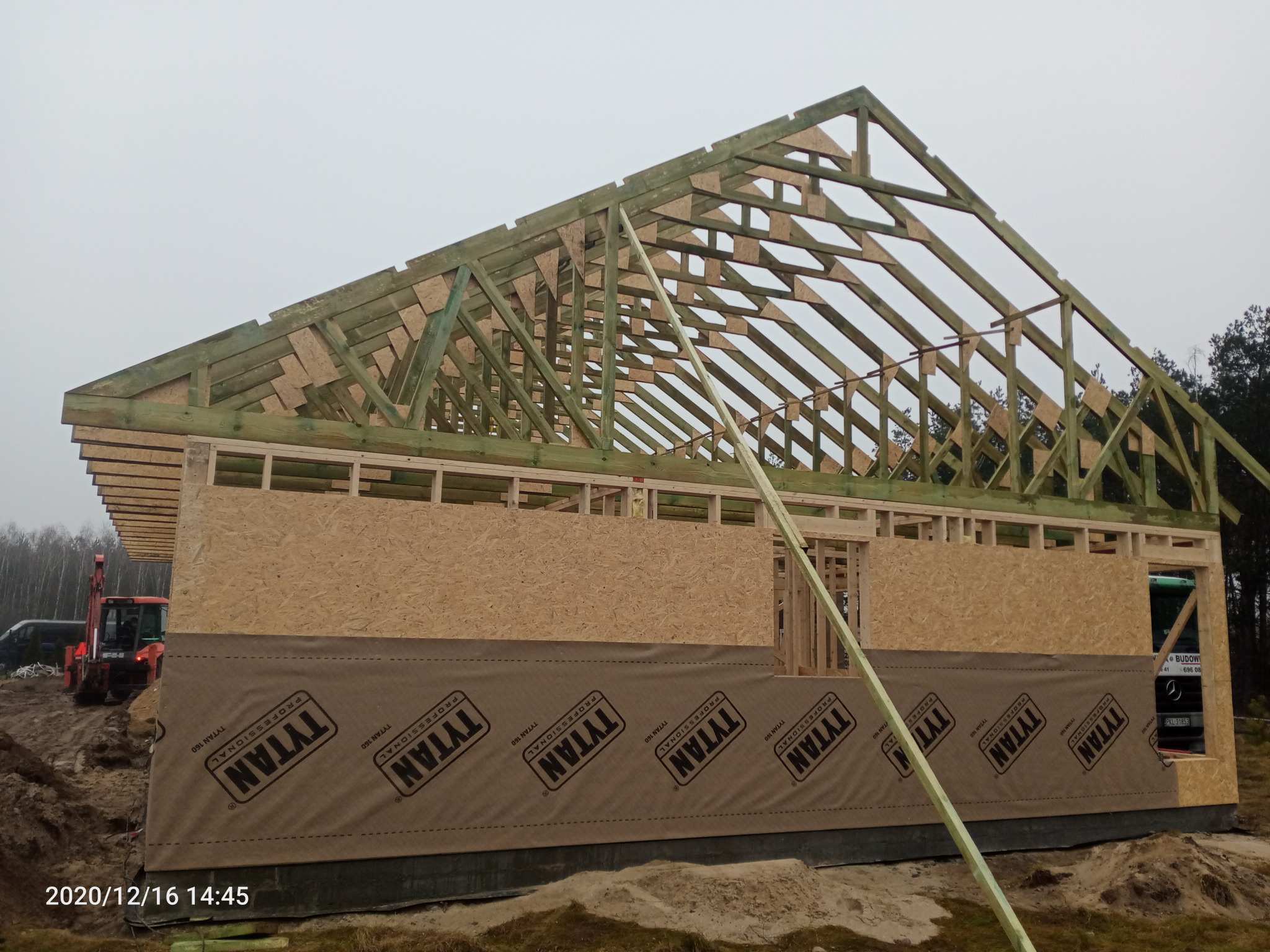 Konstrukcja szkieletowa domu w trakcie budowy, widoczne drewniane elementy konstrukcyjne i płyty OSB, zabezpieczone folią izolacyjną z nadrukowanym logo firmy TYTAN, koparka w tle.