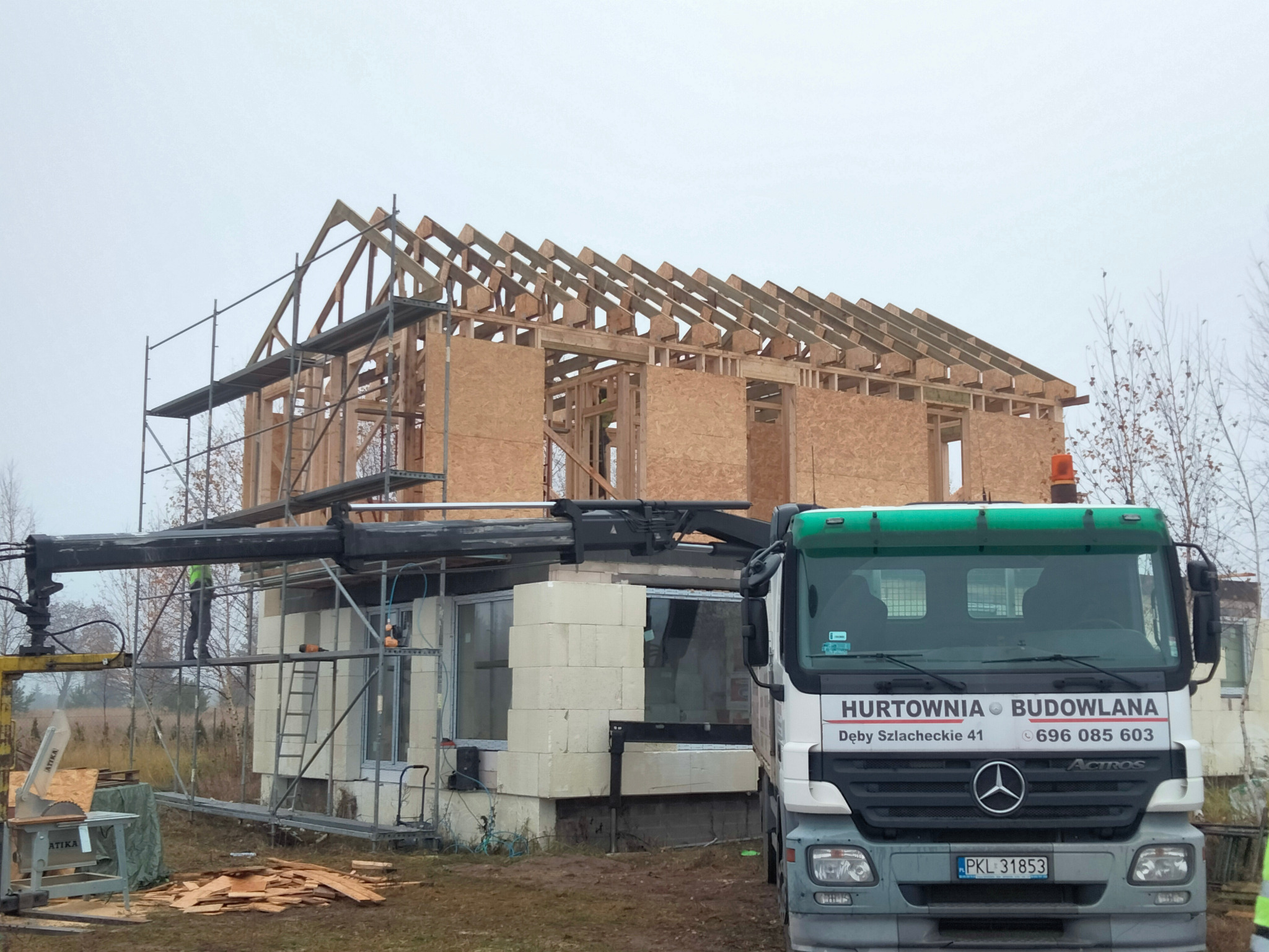 Wznoszenie konstrukcji domu szkieletowego z widocznym drewnianym szkieletem, płytami OSB, rusztowaniami, dźwigiem i zaparkowaną ciężarówką z napisem 'Hurtownia Budowlana' na tle pochmurnego nieba.