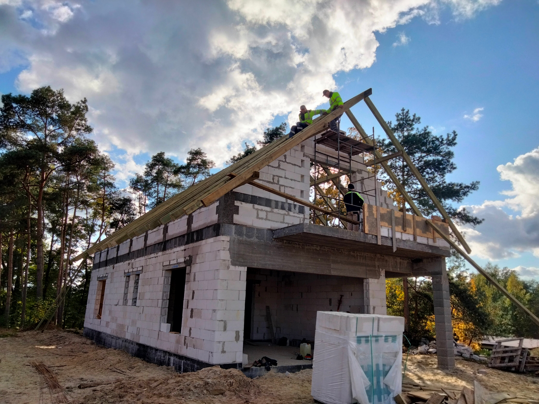 Wznoszenie budynku z białych bloczków, widoczni pracownicy na dachu montujący konstrukcję drewnianą, w tle las i błękitne niebo z chmurami.