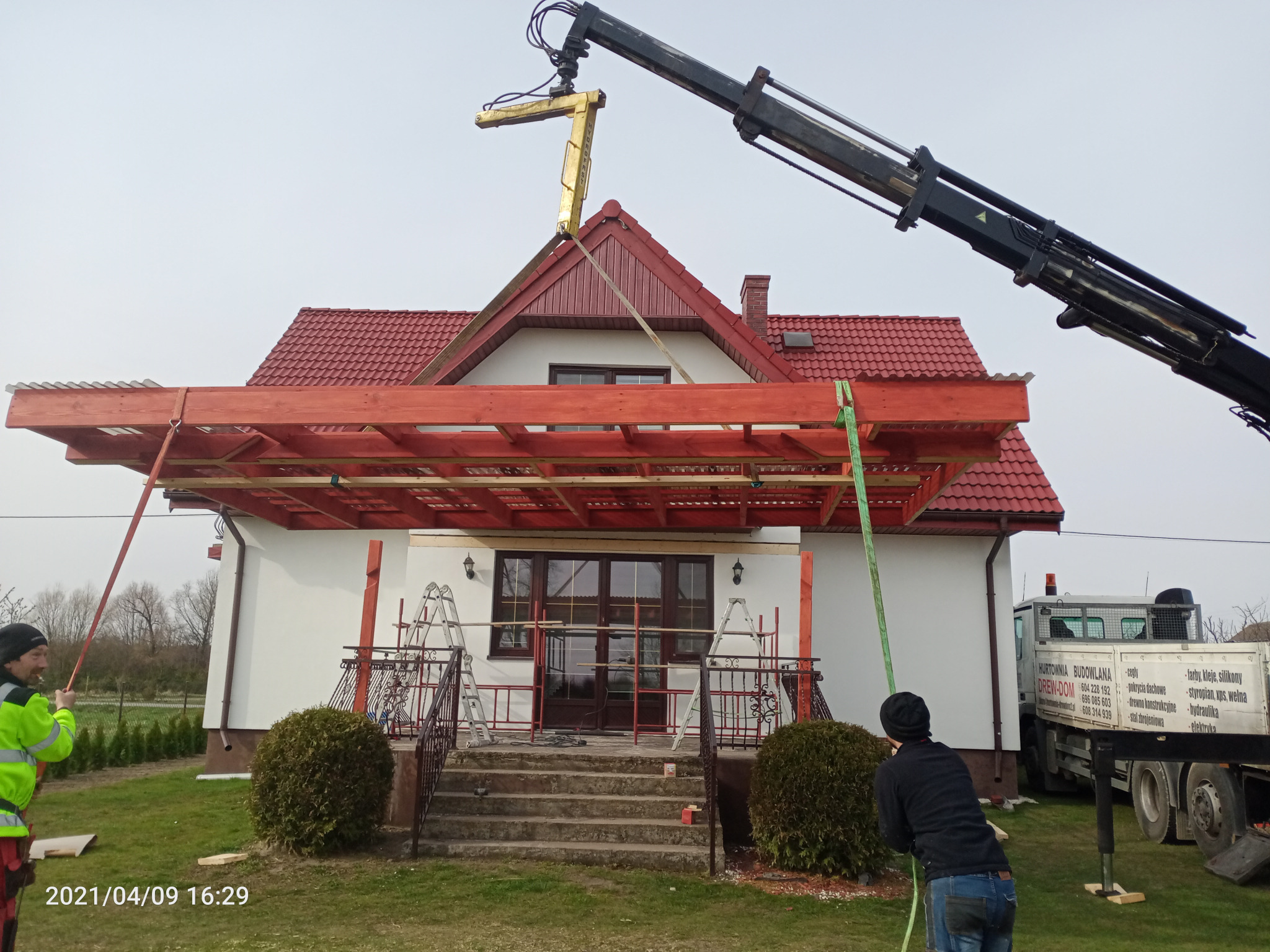 Montaż drewnianej pergoli przy domu jednorodzinnym z użyciem dźwigu, widoczni pracownicy w odzieży roboczej, elementy konstrukcyjne w kolorze czerwonym.