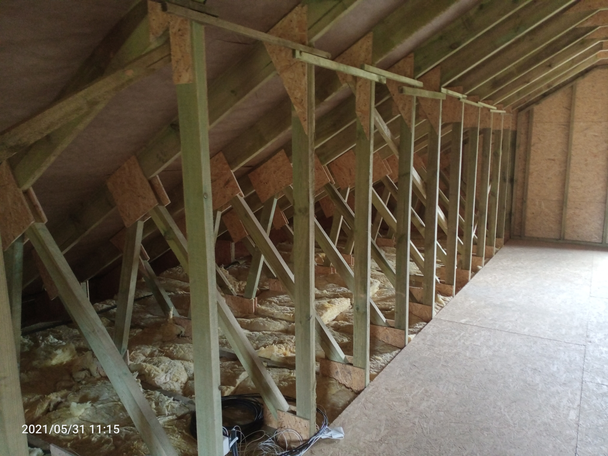 Wnętrze poddasza z drewnianą konstrukcją szkieletową i częściowo ułożoną izolacją z wełny mineralnej, widoczne przewody elektryczne w rogu podłogi z płyt OSB.