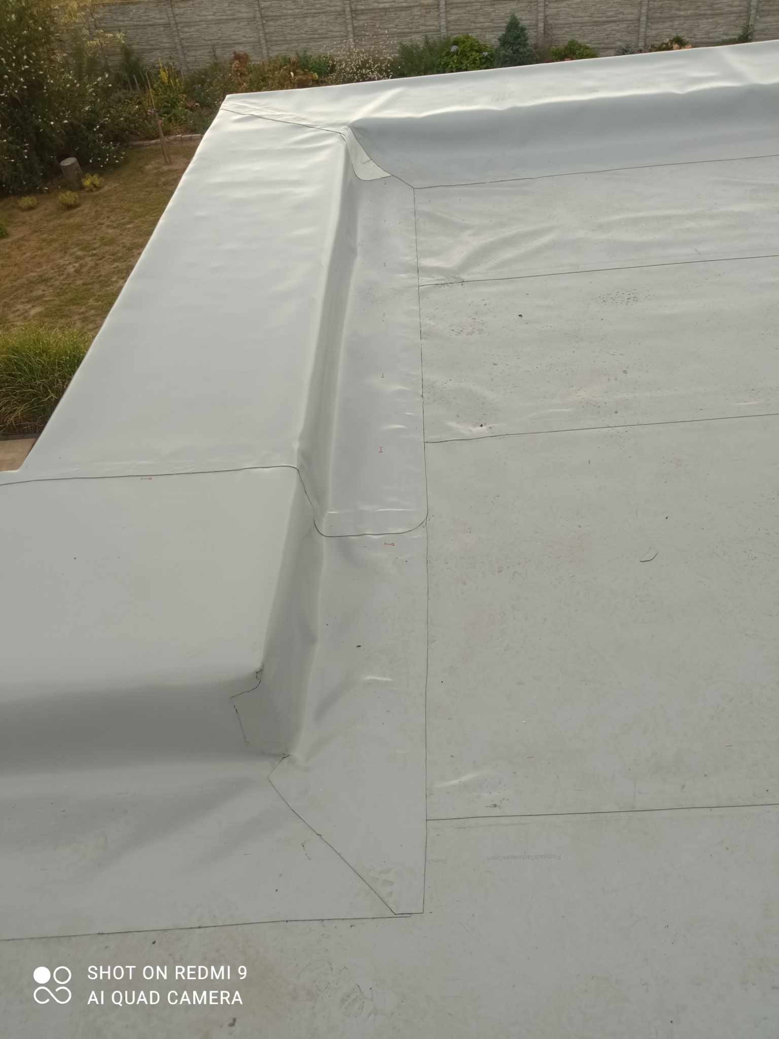 Szare, płaskie pokrycie dachu z widocznymi łączeniami i detalami obróbki blacharskiej, z fragmentem ogrodu i ogrodzenia w tle.