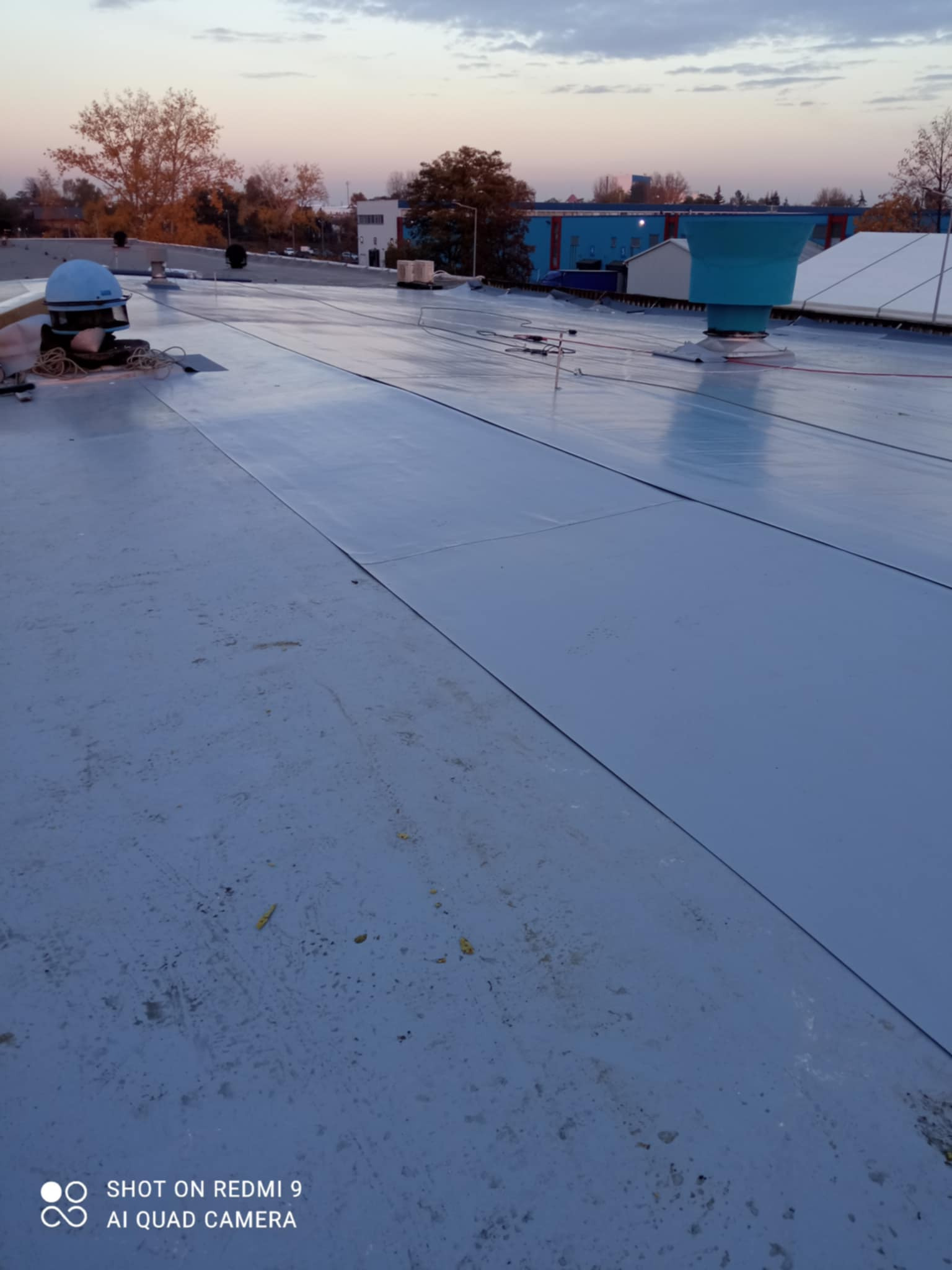 Szczelnie pokryty, płaski dach z jasnoszarej membrany bitumicznej, widoczne detale łączeń, wentylatory dachowe i elementy mocujące, w tle budynki i drzewa o zmierzchu.