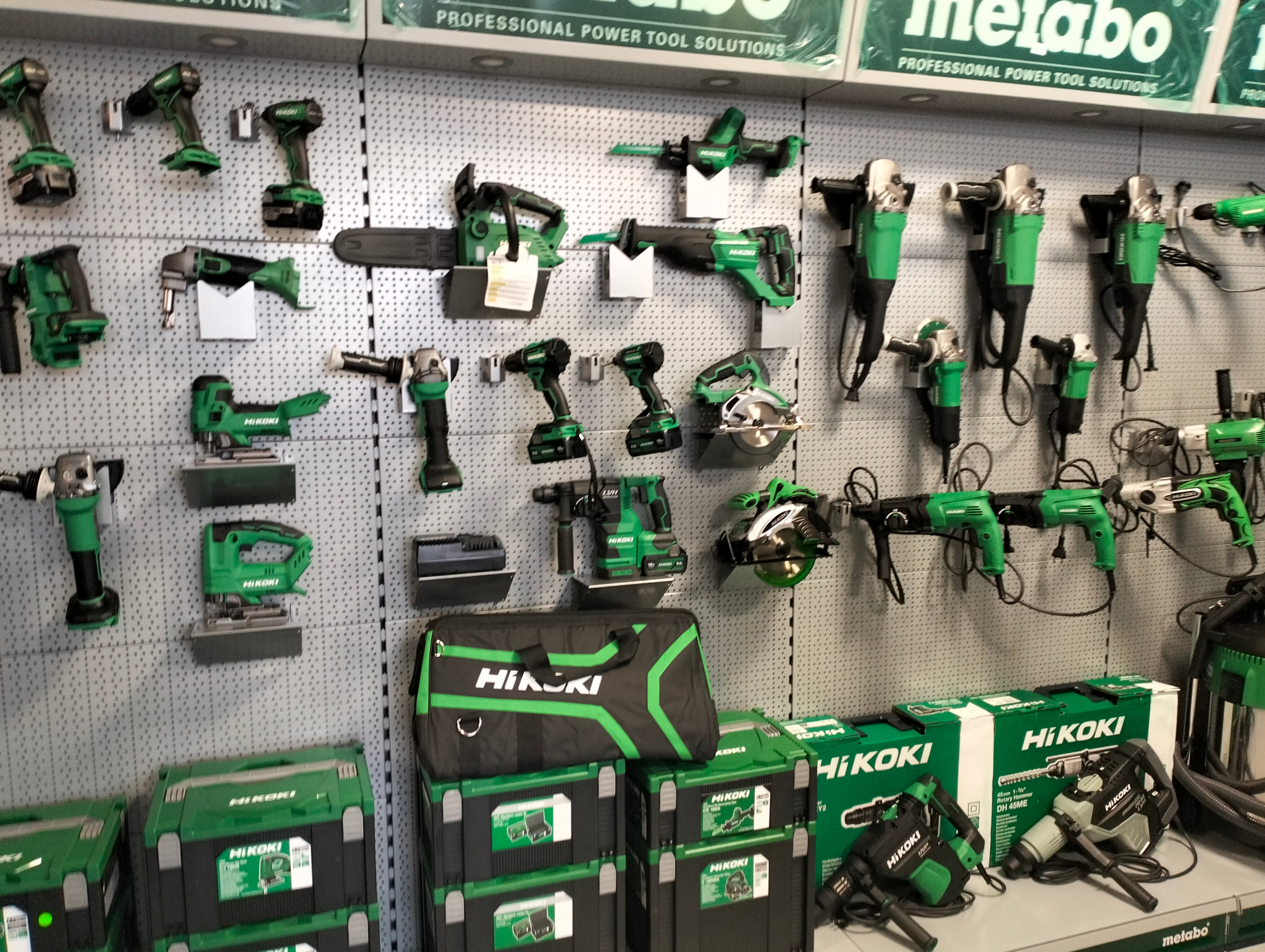 Ekspozycja różnorodnych elektronarzędzi marki HiKOKI w sklepie: wiertarki, piły, szlifierki kątowe i inne, wyeksponowane na perforowanej ścianie i w skrzynkach na dole.