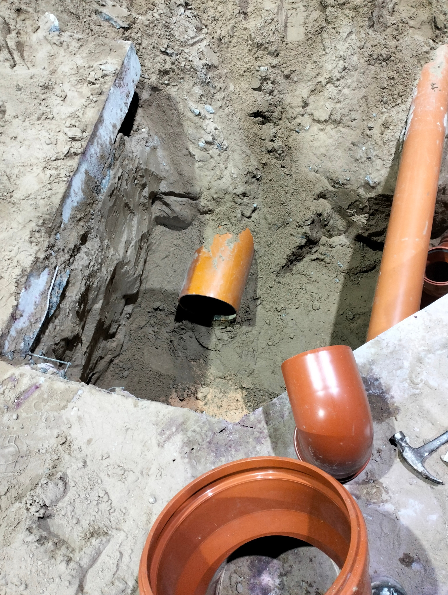 Wykop z widocznymi pomarańczowymi rurami kanalizacyjnymi, fragmentem betonowej płyty i młotkiem w tle.