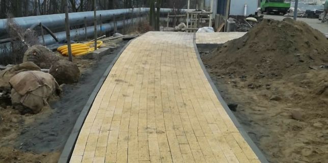 Zakrzywiony chodnik z jasnej kostki brukowej z szarą obwódką, obok przygotowane drzewka w jutowych workach i stosy ziemi.