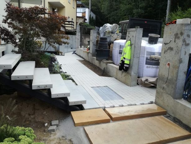 Widok na ścieżkę z jasnej kostki brukowej, schody z betonowych płyt i elementy ogrodzenia w trakcie budowy, z workami z materiałem budowlanym i odblaskową kurtką w tle.