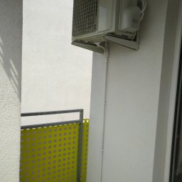 Oskar Klimatyzacja - Biały klimatyzator marki AUX zamontowany na balkonie z żółtą osłoną balustrady, widoczne rury odprowadzające skropliny.