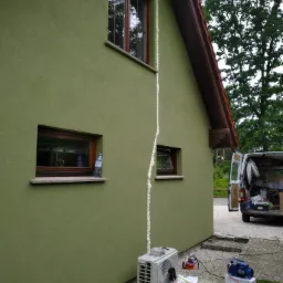 Zewnętrzna jednostka klimatyzacji zamontowana przy ścianie domu z widocznymi przewodami i urządzeniem do serwisu, ślady pianki montażowej wzdłuż rury prowadzącej do okna, samochód serwisowy w tle.