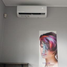 Oskar Klimatyzacja - Biały klimatyzator ścienny z wyświetlaczem temperatury 26 stopni Celsjusza, zawieszony nad plakatem reklamowym kosmetyków do włosów w salonie.