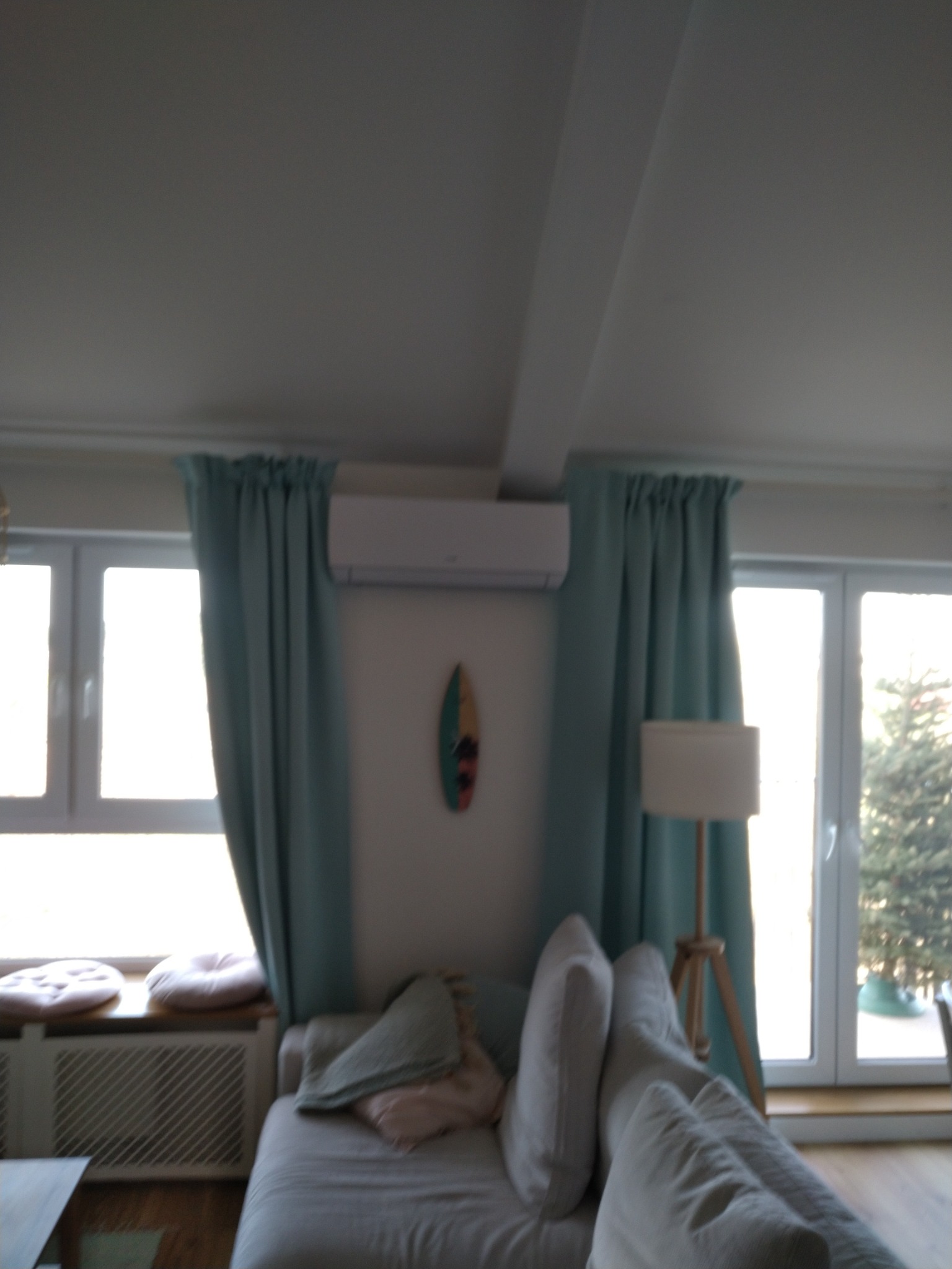 Biały klimatyzator ścienny zamontowany w jasnym salonie z turkusowymi zasłonami, dekoracyjną deską surfingową i białym narożnikiem.