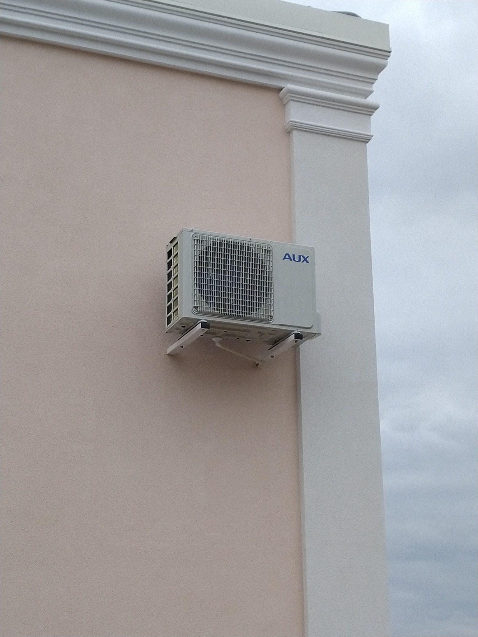 Zewnętrzna jednostka klimatyzacji marki AUX zamontowana na jasnoróżowej ścianie budynku z dekoracyjnym, białym narożnikiem.
