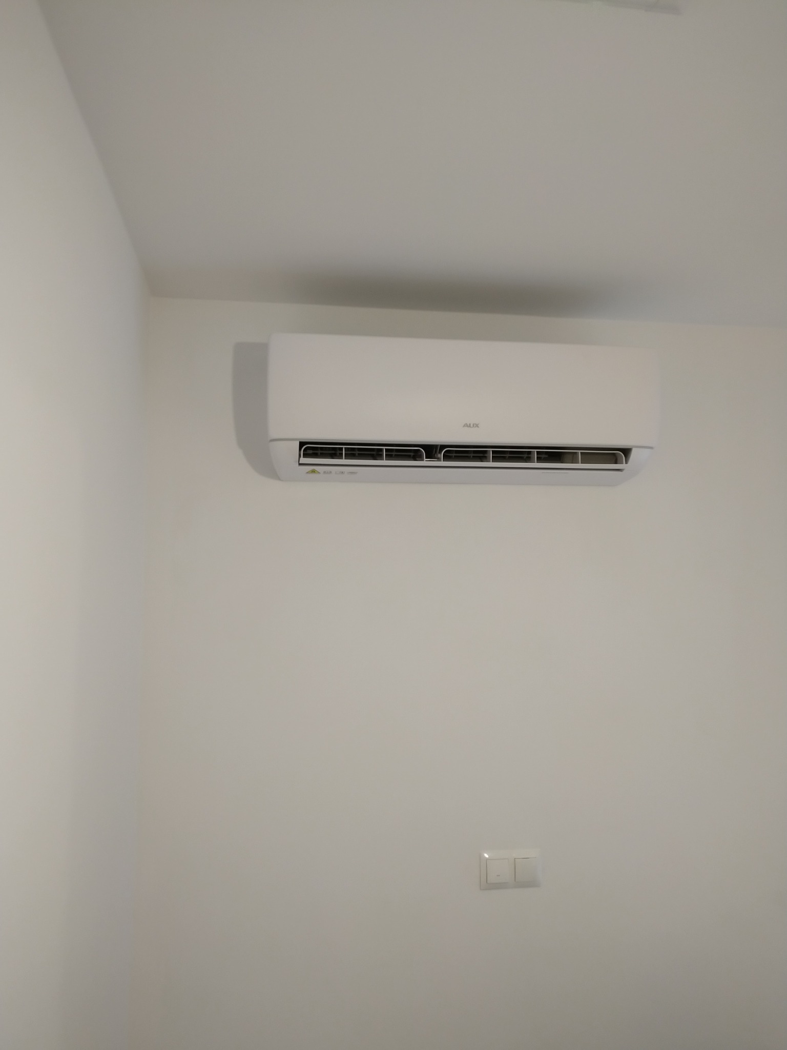 Biała jednostka klimatyzacyjna AUX zamontowana na jasnej ścianie w pomieszczeniu, widoczny przełącznik światła pod klimatyzatorem.
