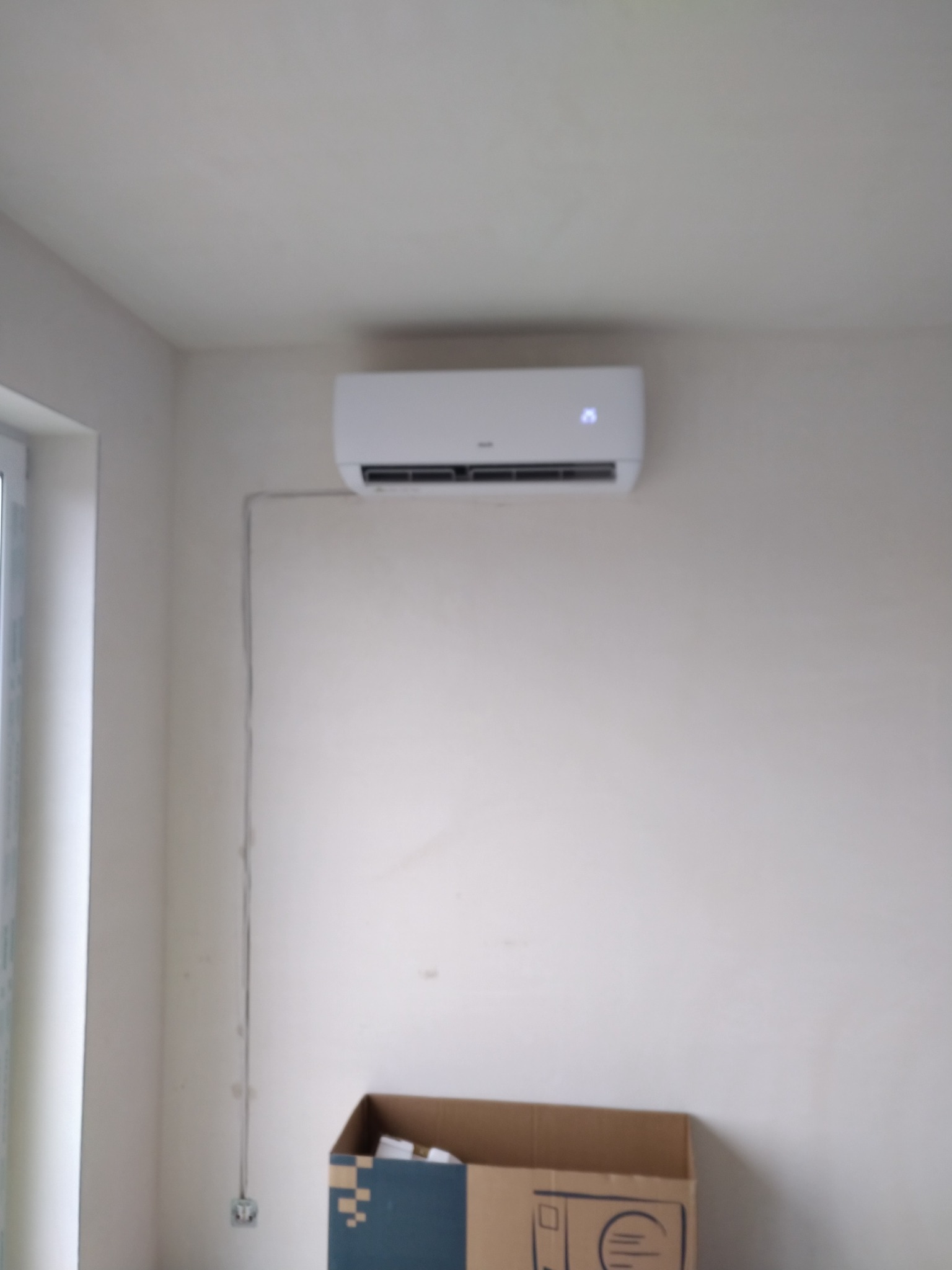 Biały klimatyzator ścienny zamontowany w jasnym pomieszczeniu z widocznymi przewodami i kartonem na podłodze.
