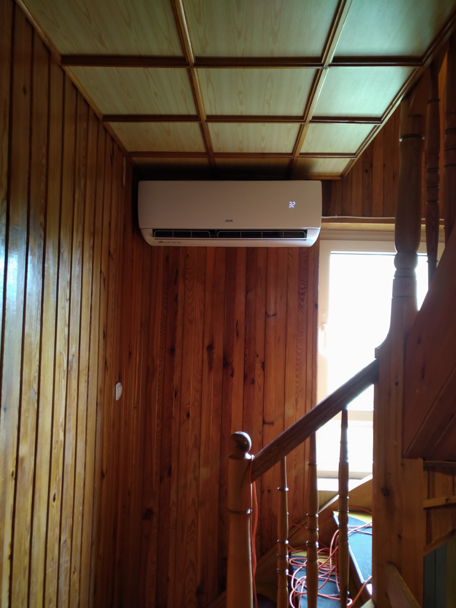 Biały klimatyzator AUX zamontowany na drewnianej ścianie w pomieszczeniu z drewnianym sufitem kasetonowym, widoczne schody z drewnianą balustradą.