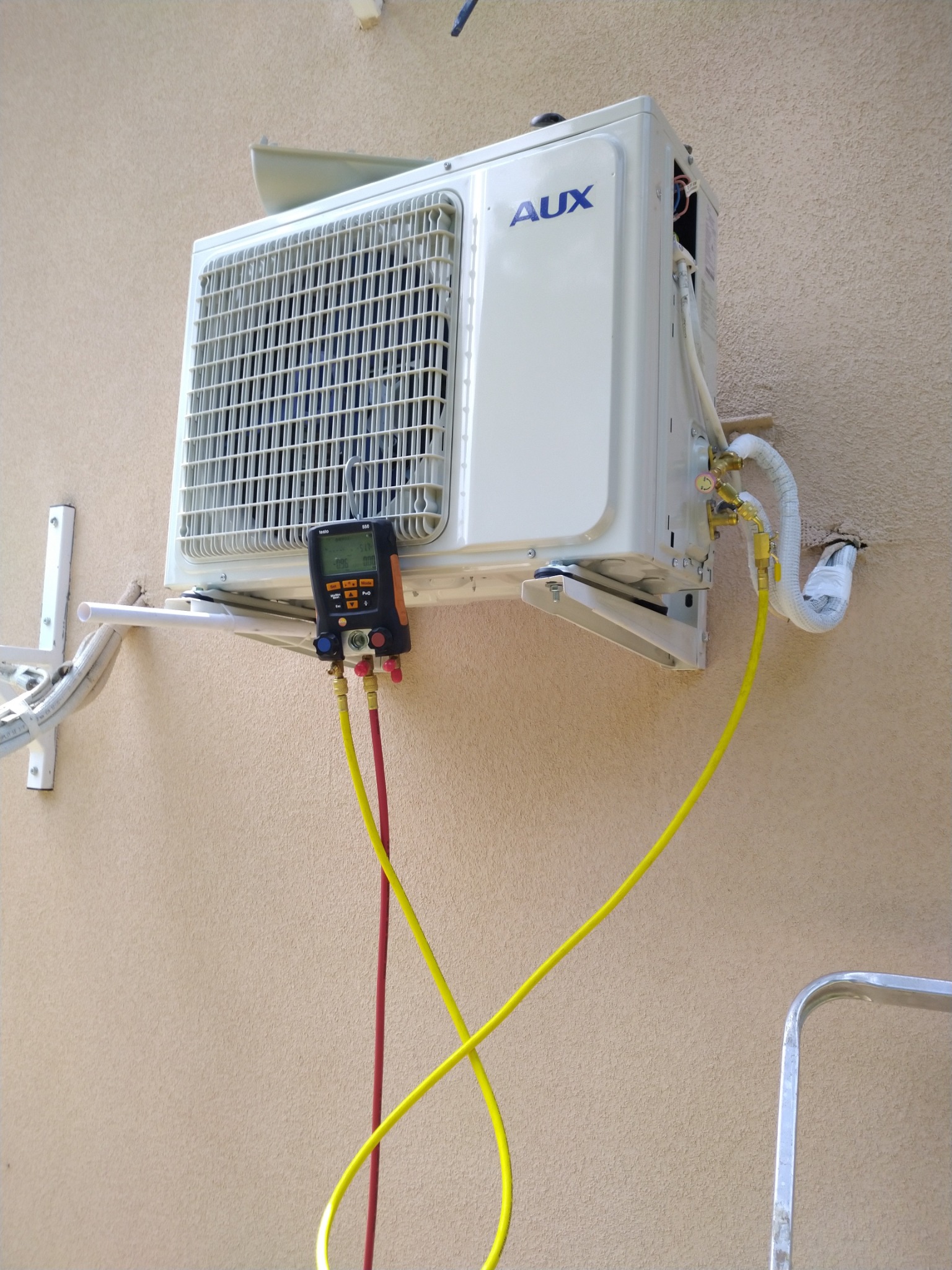 Biała jednostka zewnętrzna klimatyzacji AUX zamontowana na ścianie budynku, podłączona do manometru cyfrowego za pomocą żółtych i czerwonych przewodów serwisowych.
