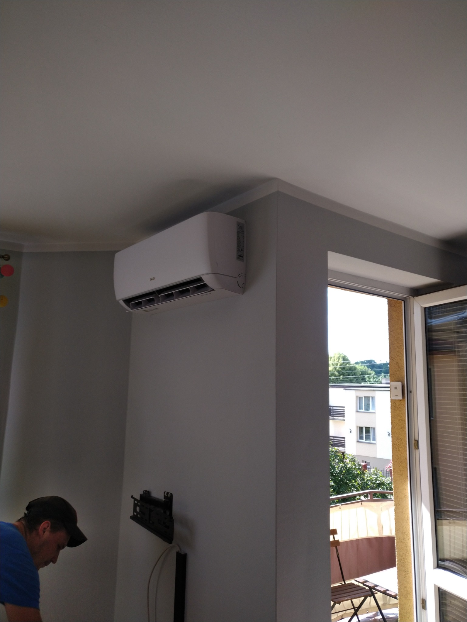 Biały klimatyzator AUX zamontowany na szarej ścianie w pokoju z wyjściem na balkon, widoczny fragment instalatora z boku.