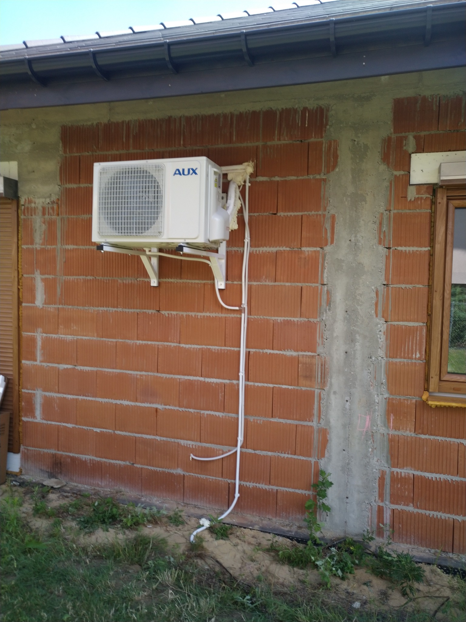 Biała jednostka zewnętrzna klimatyzacji marki AUX zamontowana na ceglanej ścianie budynku w trakcie instalacji, widoczne rury odprowadzające skropliny i przewody elektryczne.