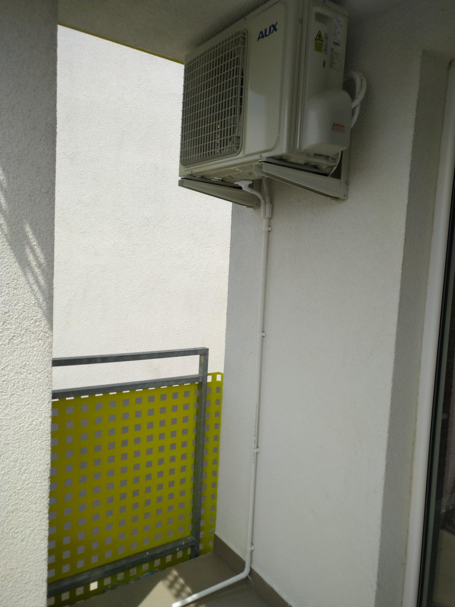 Biały klimatyzator marki AUX zamontowany na balkonie z żółtą osłoną balustrady, widoczne rury odprowadzające skropliny.