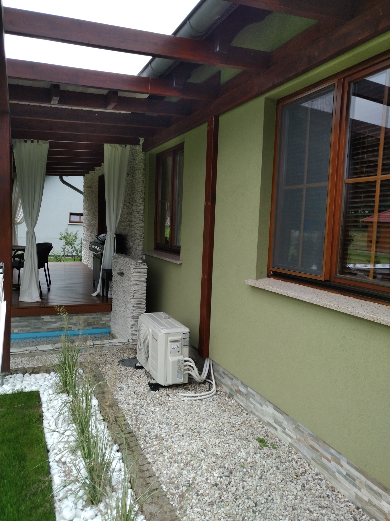 Biała jednostka zewnętrzna klimatyzacji zamontowana na żwirowym podłożu przy zielonej ścianie domu z drewnianymi oknami, widoczna część tarasu z meblami i zasłonami.