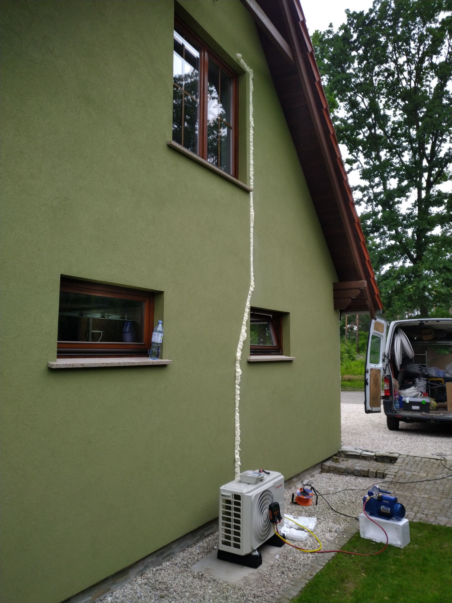 Zewnętrzna jednostka klimatyzacji zamontowana przy ścianie domu z widocznymi przewodami i urządzeniem do serwisu, ślady pianki montażowej wzdłuż rury prowadzącej do okna, samochód serwisowy w tle.