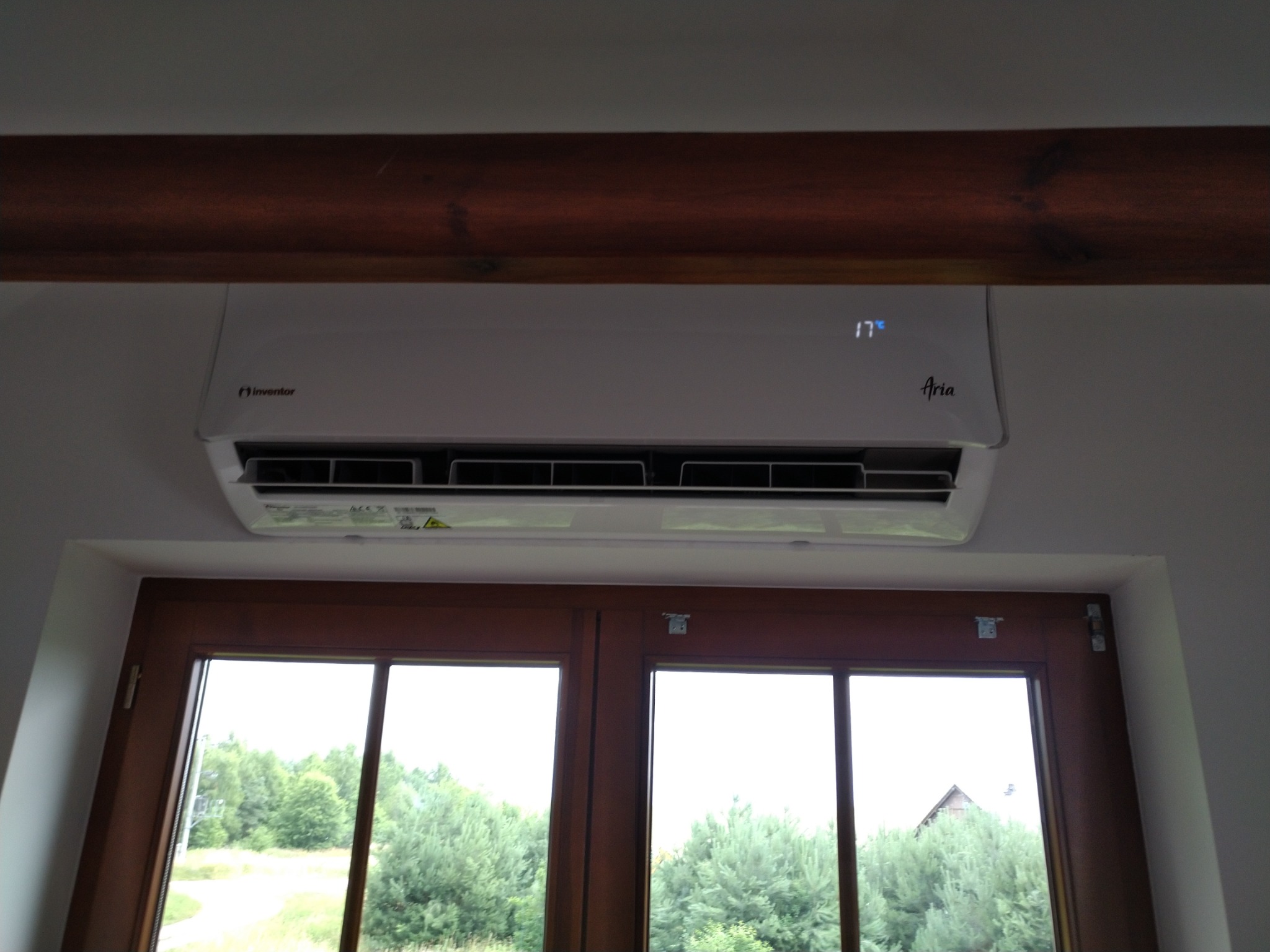 Biały klimatyzator Inverter Aria zamontowany nad oknem z widokiem na zieleń, temperatura ustawiona na 17 stopni Celsjusza, widoczna drewniana belka stropowa.