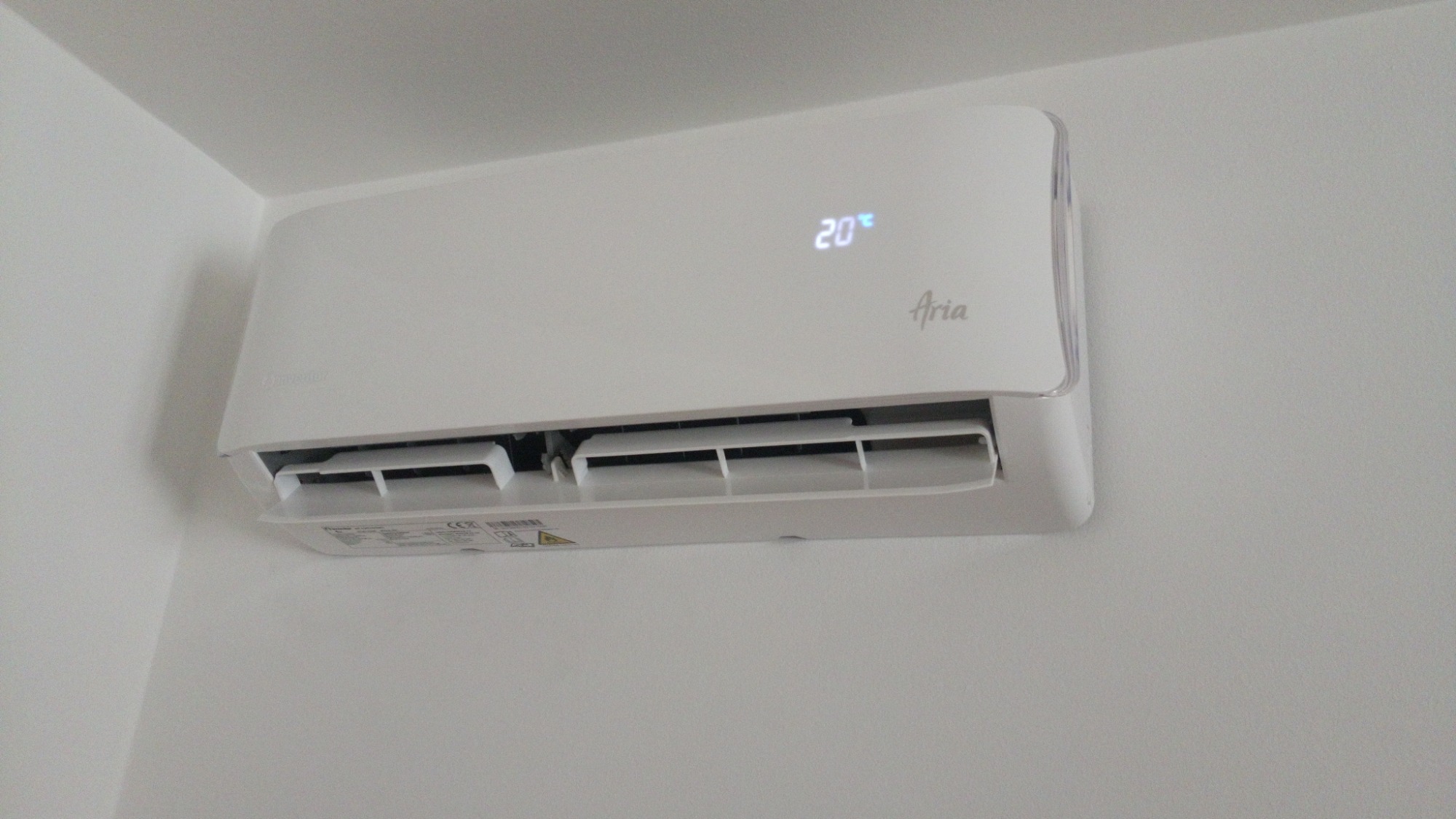 Biały klimatyzator Aria z wyświetlaną temperaturą 20 stopni Celsjusza, zamontowany na białej ścianie w rogu pomieszczenia.