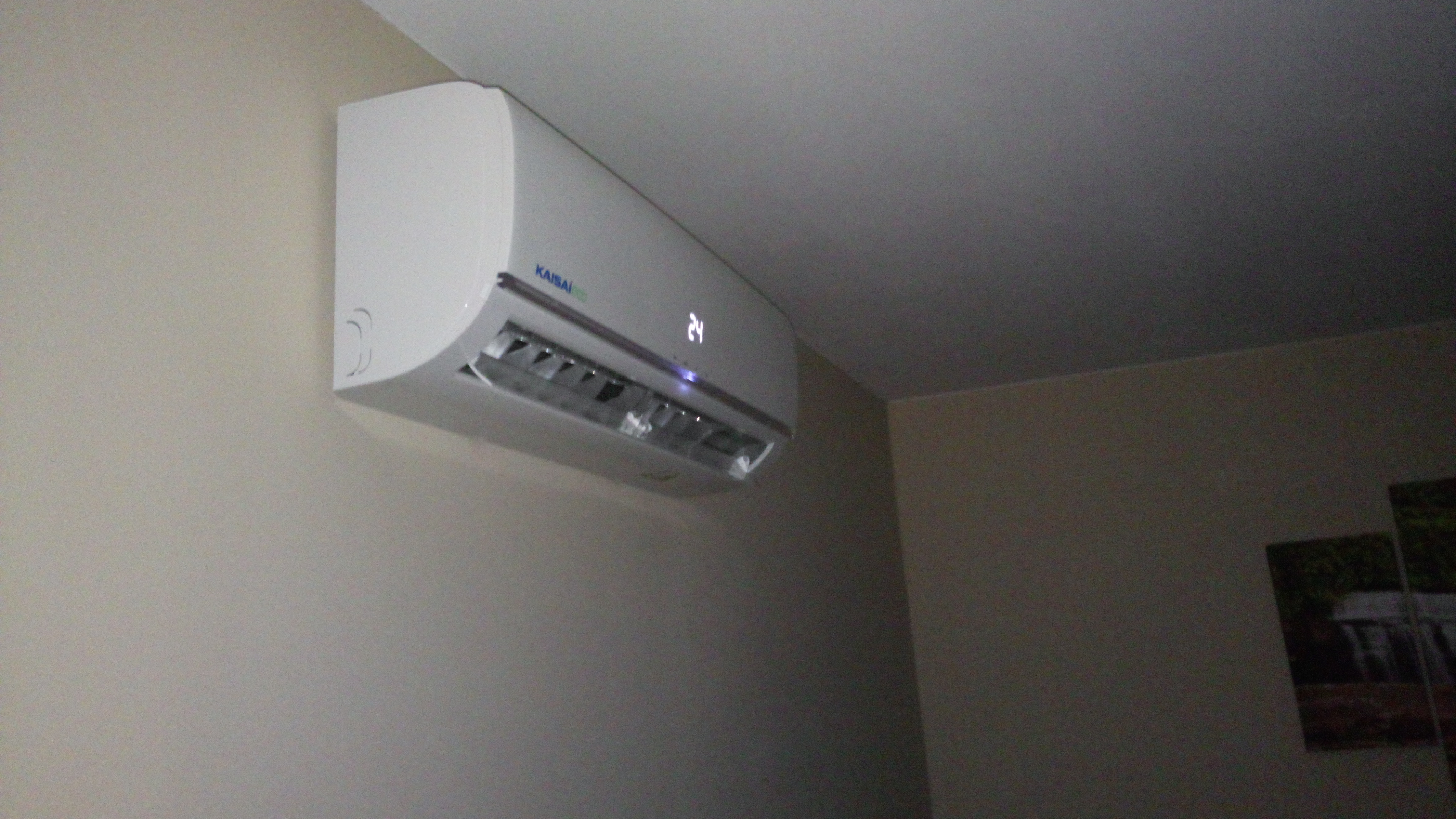Biała jednostka klimatyzacji marki Kaisai zamontowana na ścianie w pomieszczeniu, wyświetlająca temperaturę 24 stopnie Celsjusza, z widocznym fragmentem obrazu na ścianie w tle.