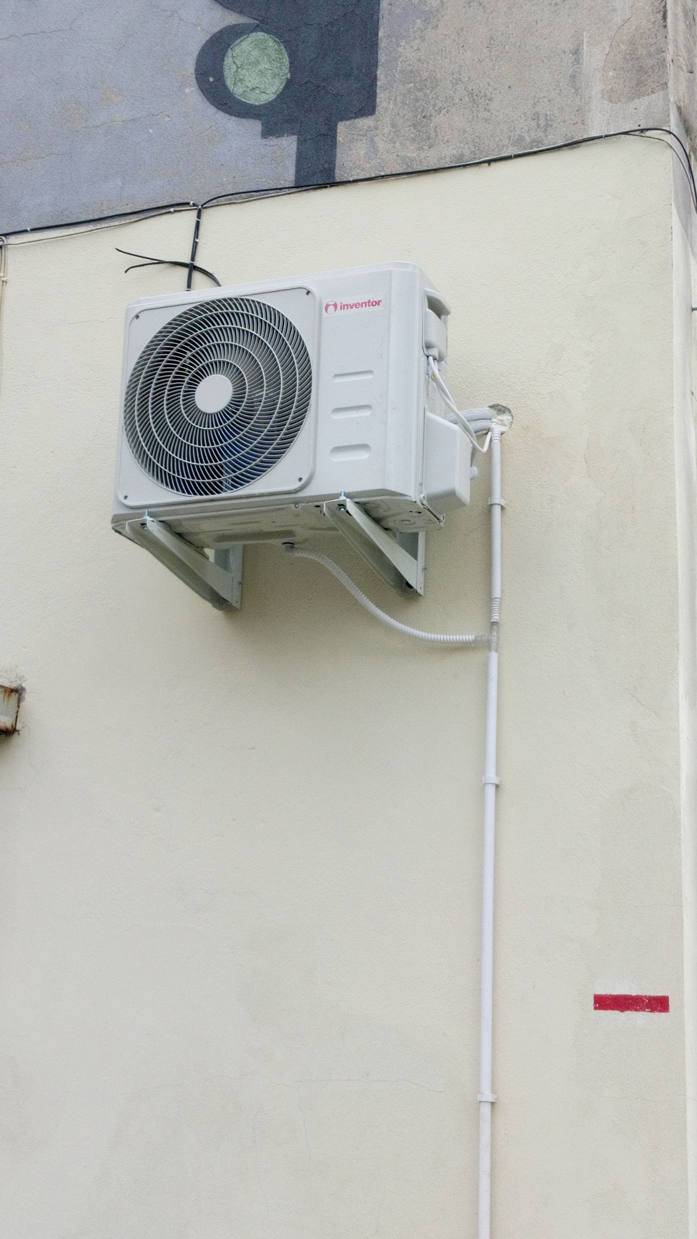 Zewnętrzna jednostka klimatyzatora marki Inventor zamontowana na kremowej ścianie budynku, widoczne rury odprowadzające skropliny i przewody elektryczne, na ścianie ślady po malowaniu.