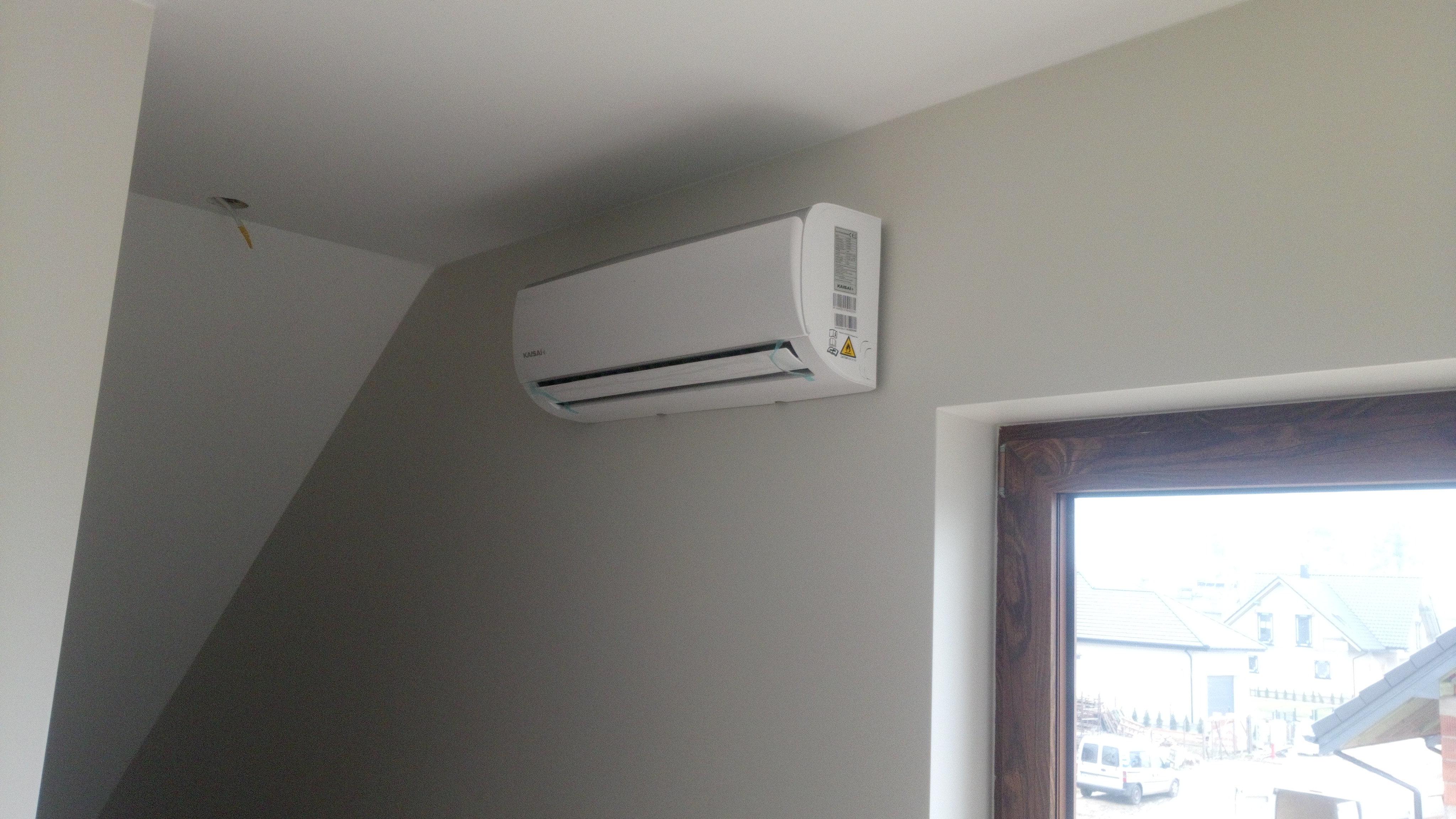 Biały klimatyzator marki Kaisai zamontowany na szarej ścianie w pomieszczeniu z oknem o drewnianej ramie, widok na budynki z zewnątrz.