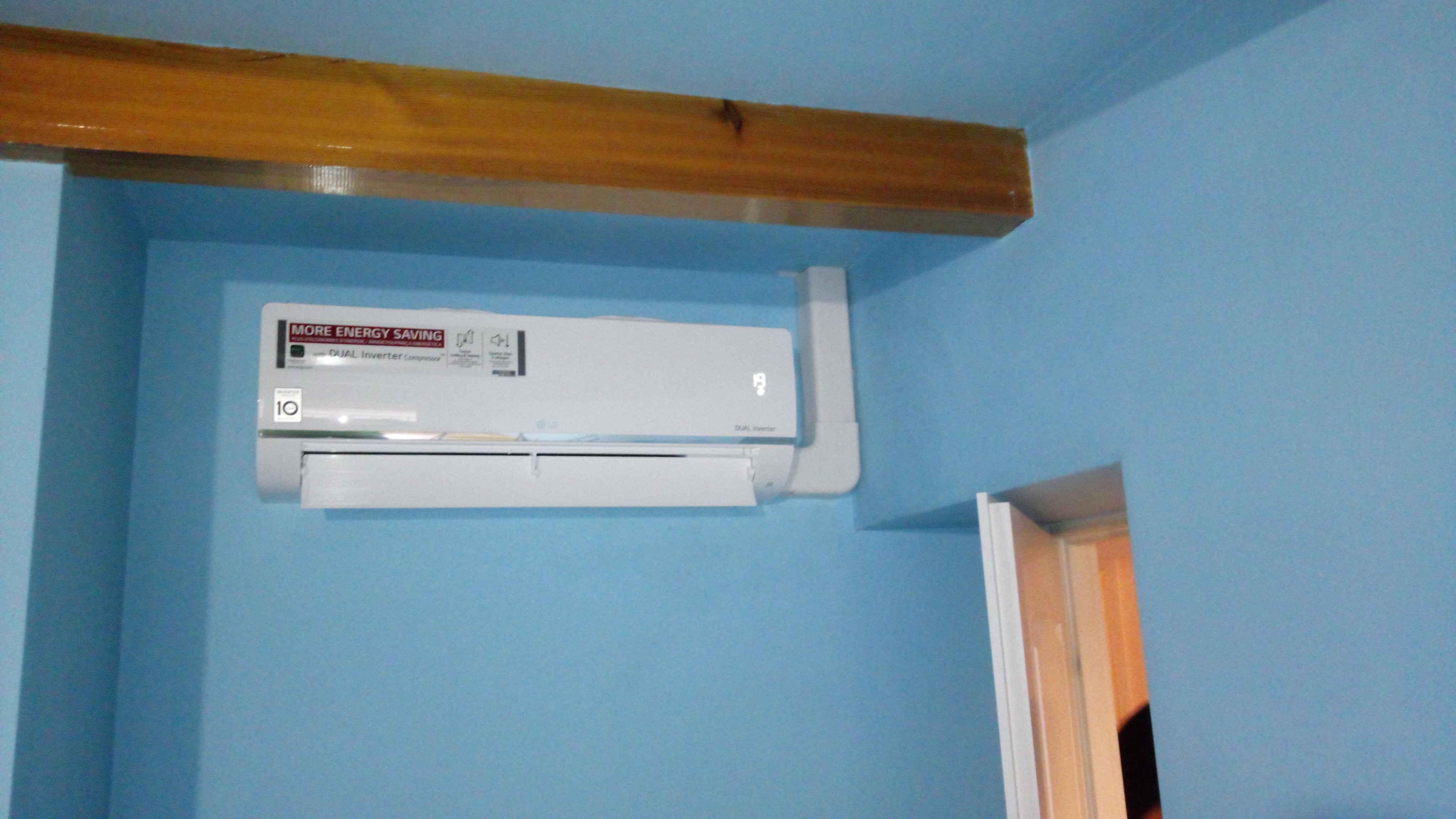 Biały klimatyzator LG z wyświetlaczem temperatury, zamontowany na błękitnej ścianie w pokoju z drewnianą belką stropową i fragmentem futryny drzwi.