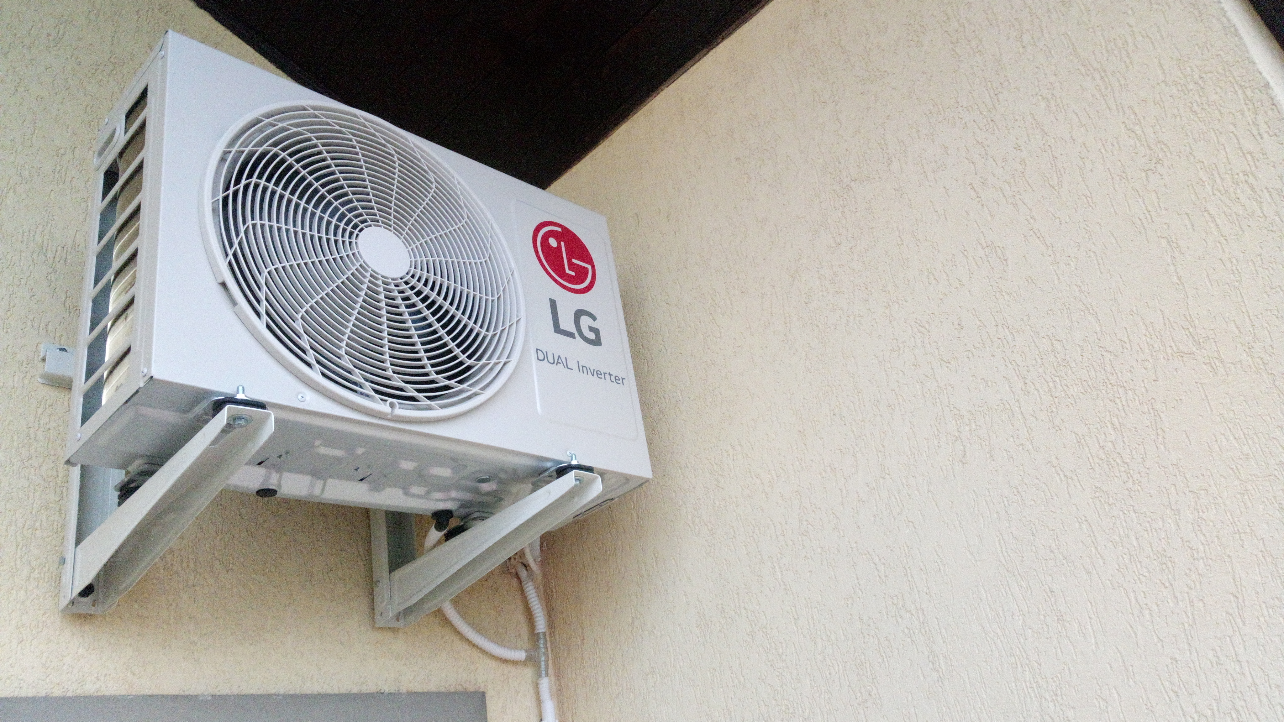 Zewnętrzna jednostka klimatyzatora LG Dual Inverter zamontowana na kremowej elewacji budynku, widoczne białe wsporniki i przewody.