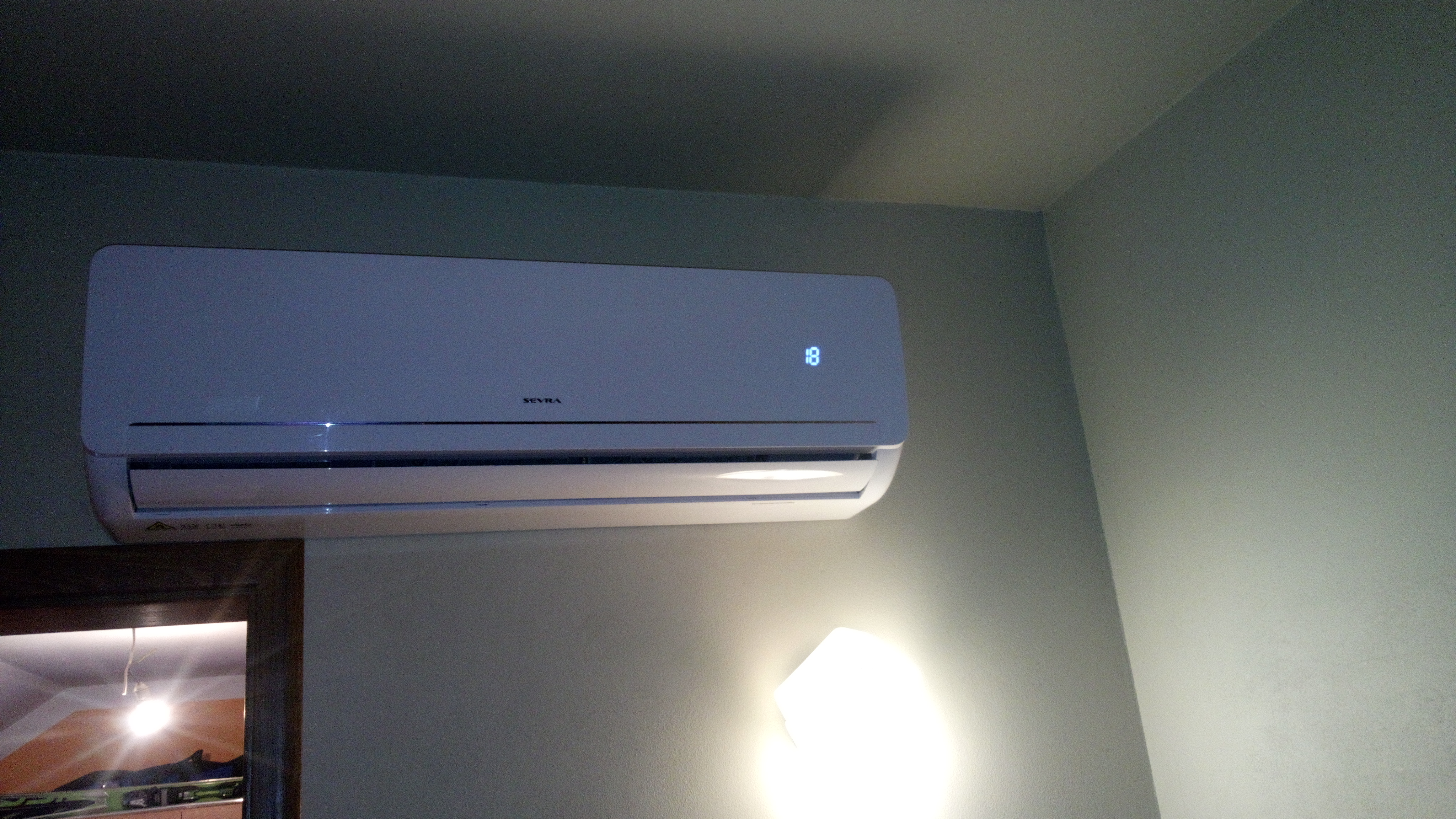 Biała jednostka wewnętrzna klimatyzacji marki Seyra zamontowana na ścianie w pokoju, z wyświetlaczem temperatury ustawionym na 18 stopni.