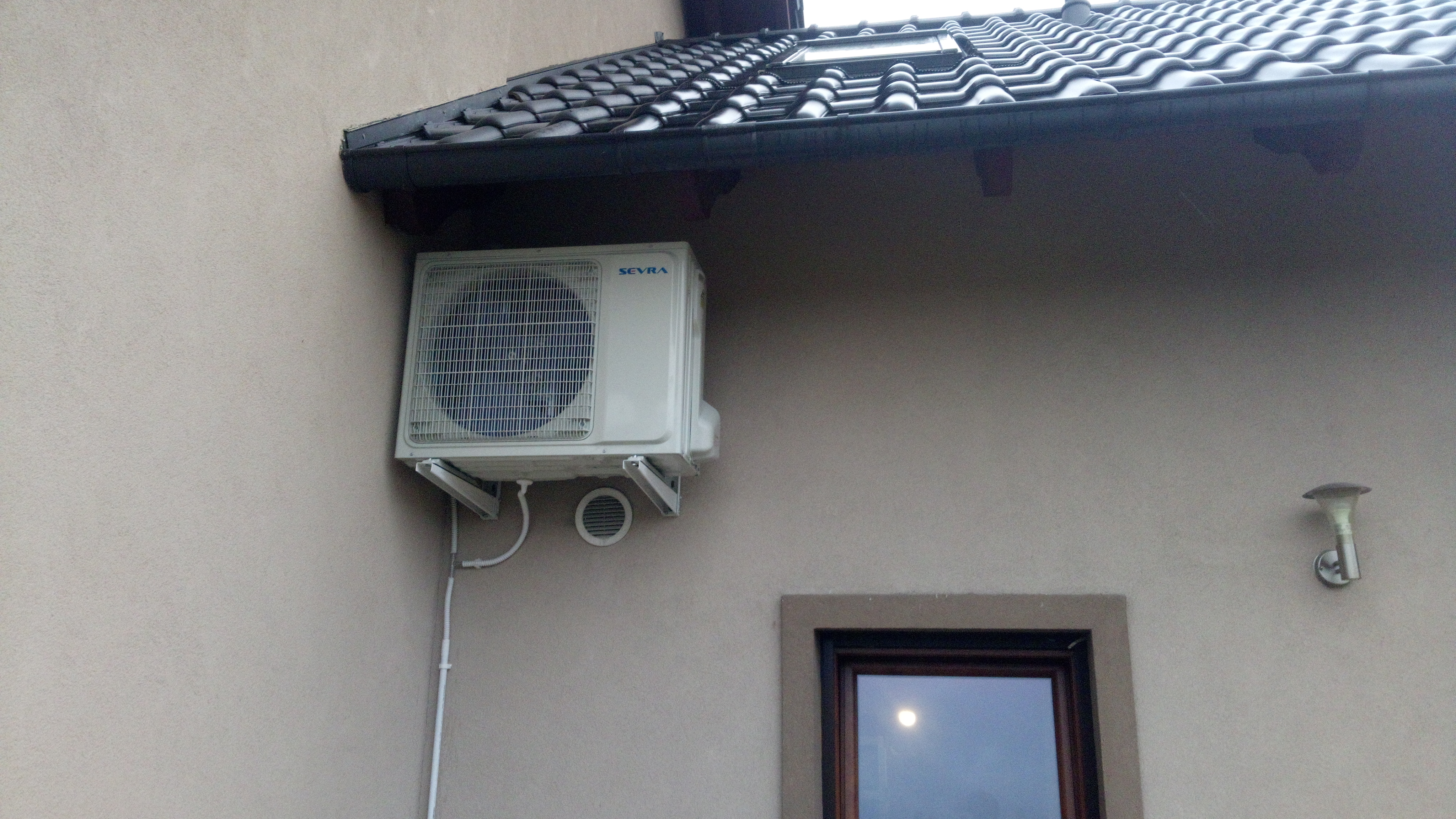 Zewnętrzna jednostka klimatyzacji marki Sevra zamontowana na elewacji domu pod okapem, widoczne rury i przewody odprowadzające skropliny.