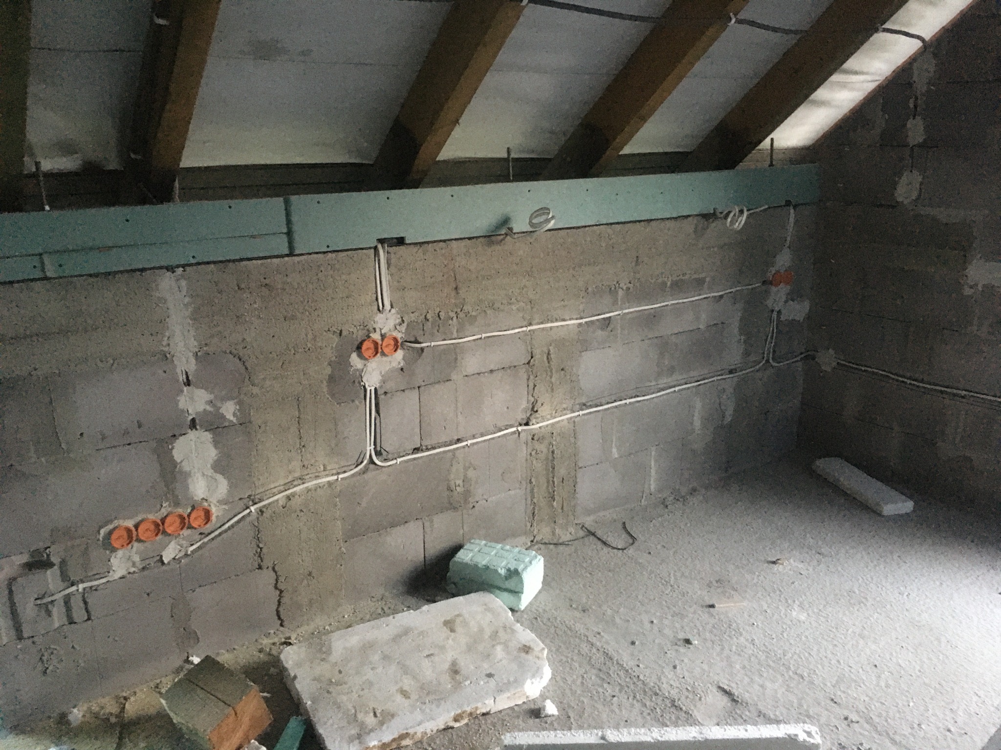 Surowy strych z instalacją elektryczną: kable w peszlach, puszki podtynkowe w ścianie z betonu komórkowego. Widoczne drewniane belki stropowe i ocieplenie.