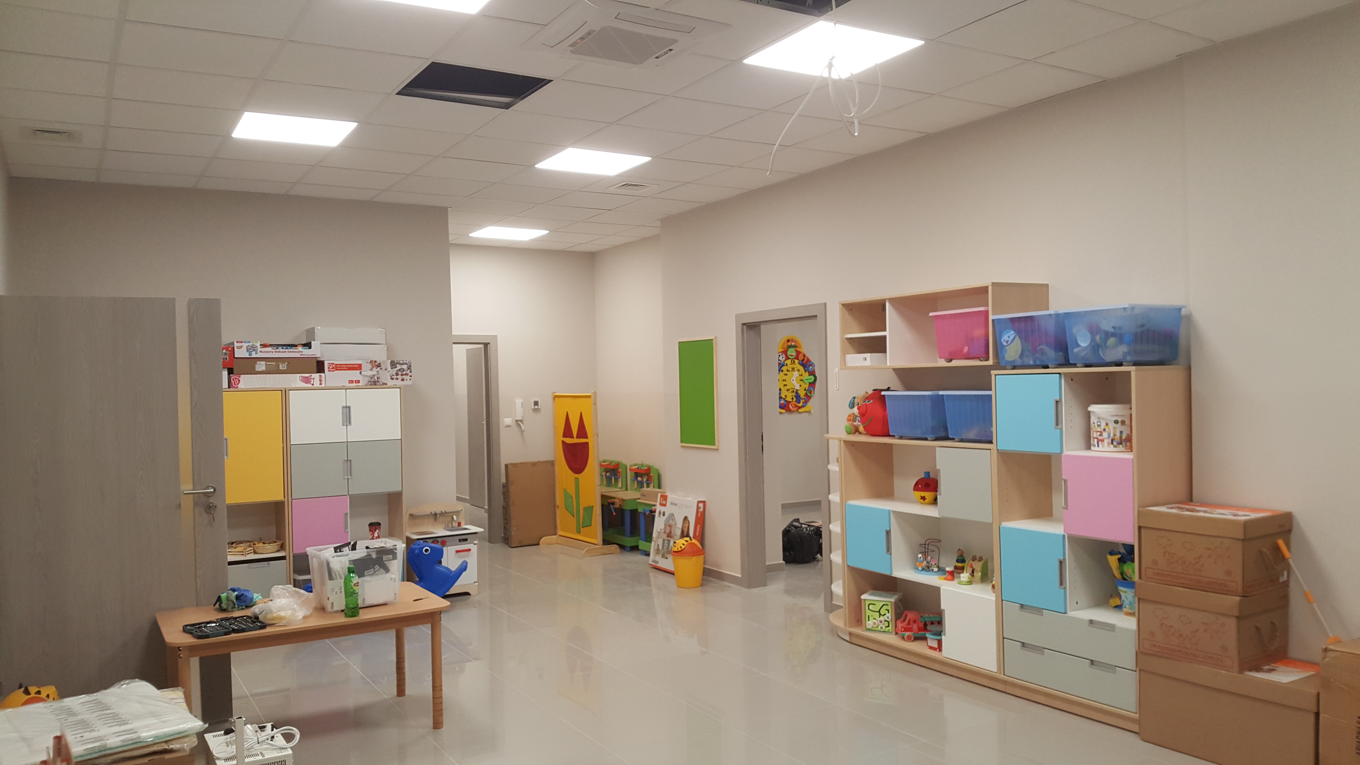 Jasne, przestronne wnętrze nowo wykończonego przedszkola z pomalowanymi ścianami, błyszczącą podłogą, kolorowymi szafkami i zabawkami; widoczne niedokończone prace przy suficie podwieszanym.