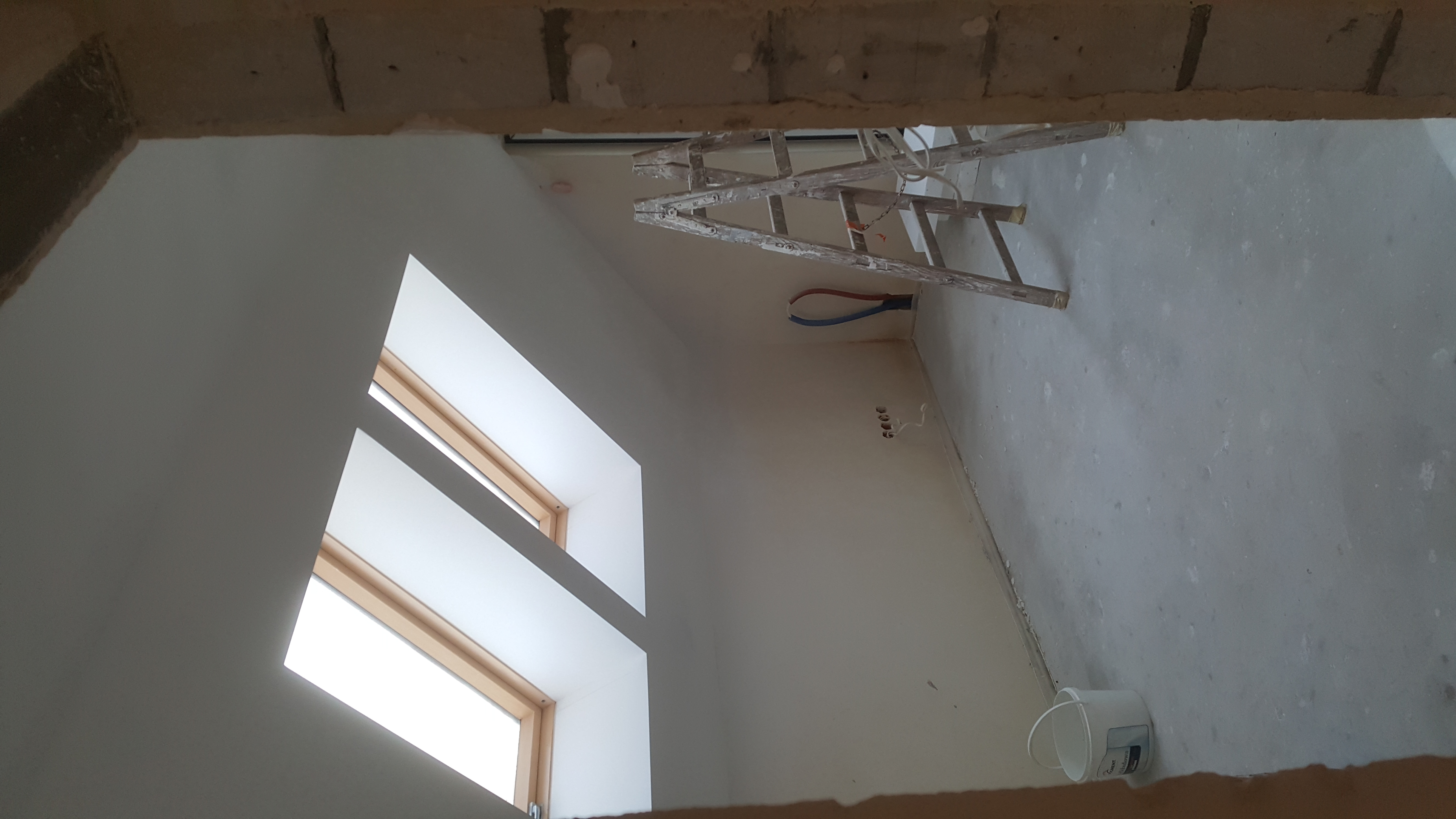 Wnętrze poddasza z oknami dachowymi w trakcie prac malarskich, widoczna drabina malarska, ściany po gładziowaniu, przygotowane do malowania, widoczne gniazdka elektryczne i rury instalacji.