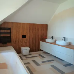 Nowoczesna łazienka na poddaszu z drewnianą ścianą, wiszącą toaletą, białą szafką z drewnianym blatem i umywalkami nablatowymi oraz grzejnikiem drabinkowym w kolorze czarnym.