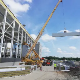 Żuraw montuje płytę warstwową na budowie hali z widoczną stalową konstrukcją szkieletową, pracownicy w podnośniku koszowym i na ziemi, oraz stosy płyt czekających na montaż.