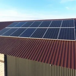 Panele słoneczne zamontowane na dachu z blachy trapezowej w kolorze ceglastym, widoczne z perspektywy lekko z góry, błękitne niebo w tle.