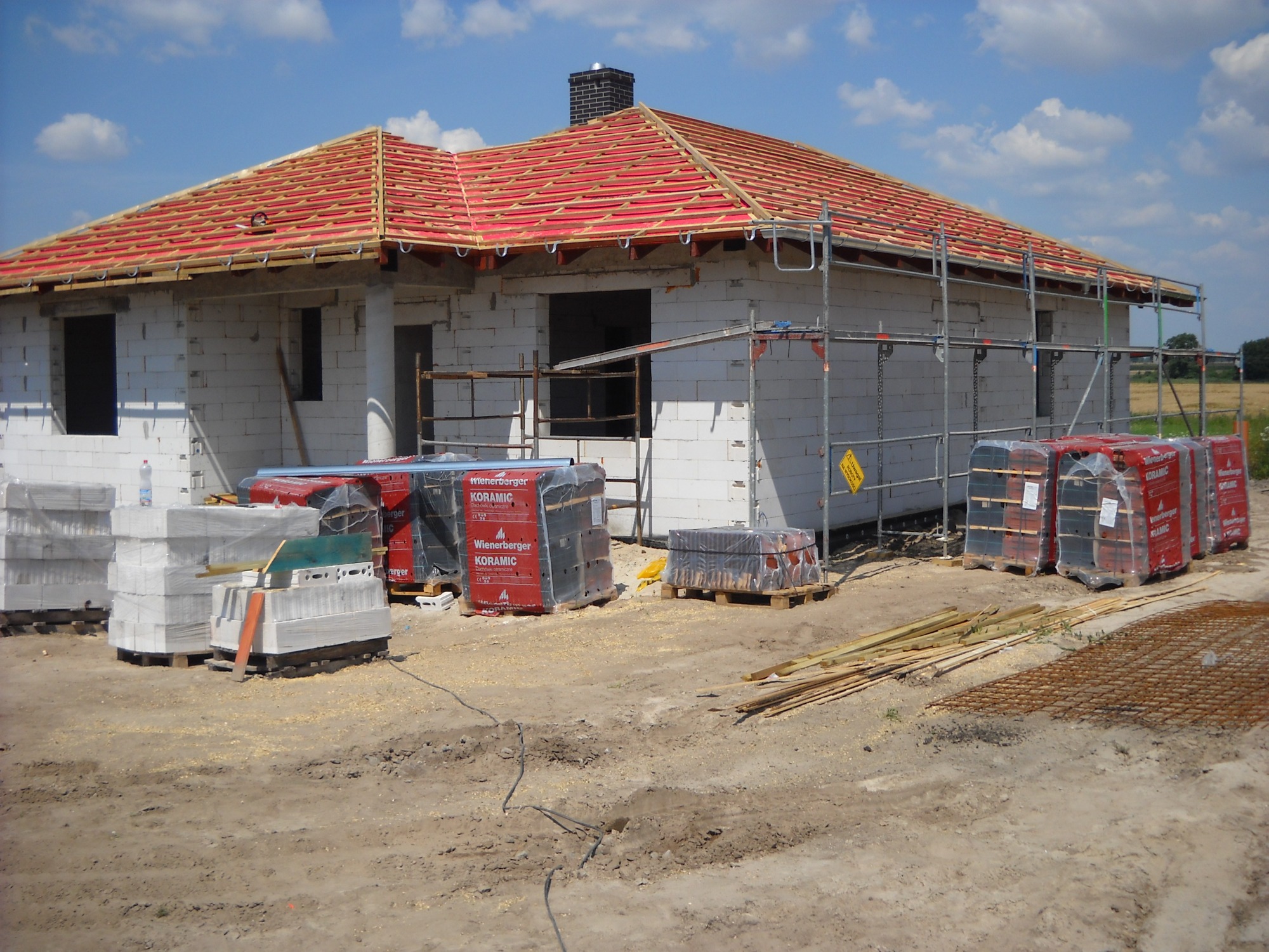 Budowa domu jednorodzinnego w trakcie montażu dachu z czerwonej dachówki ceramicznej Wienerberger Koramic, widoczne rusztowania i materiały budowlane na placu budowy.