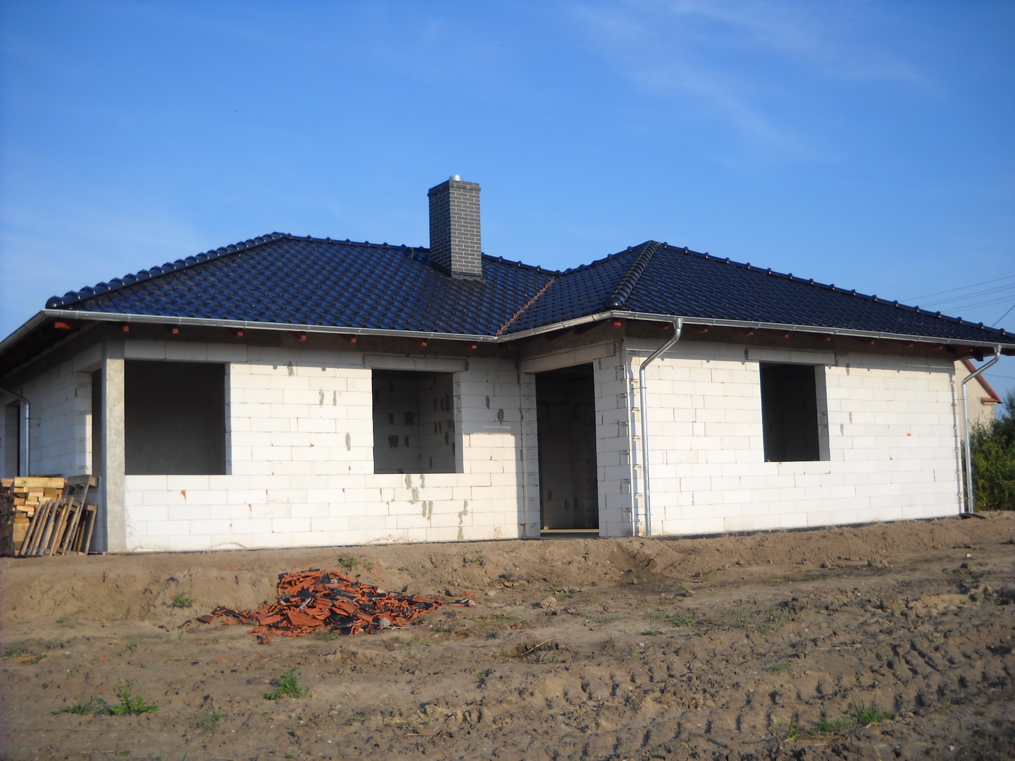 Dom w stanie surowym zamkniętym: czarne dachówki na skośnym dachu, szary komin z cegły, białe ściany z pustaków, stos drewna i sterta pomarańczowych dachówek na ziemi.