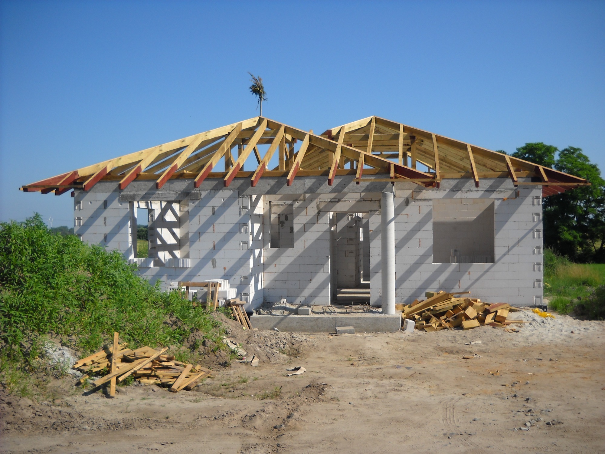 Surowy stan domu w budowie z widoczną konstrukcją dachu z drewnianych belek i ścianami z białych bloczków, na szczycie dachu umieszczono symboliczny wieniec.